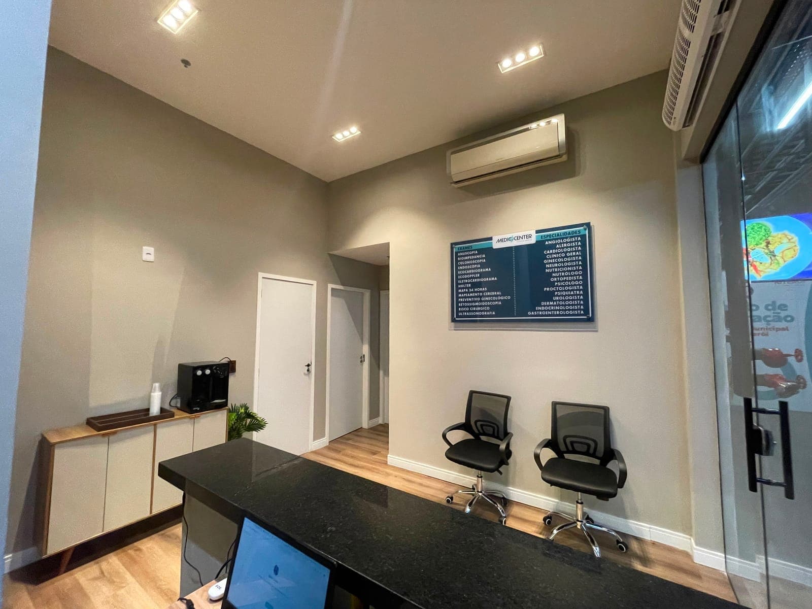Sala de espera da Mediccenter, com poltronas e o painel de exames e especialidades na parede